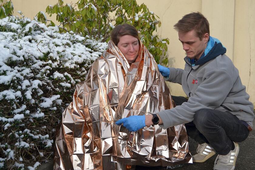 Eine Person hüllt eine andere in eine Rettungsdecke, im Hintergrund ein Strauch voll Schnee
