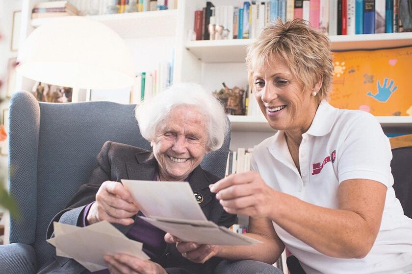 Eine Pflegerin sitzt auf der Armelehne des Sessels und schaut sich mit der Seniorin im Sessel einen Beipackzettel an.