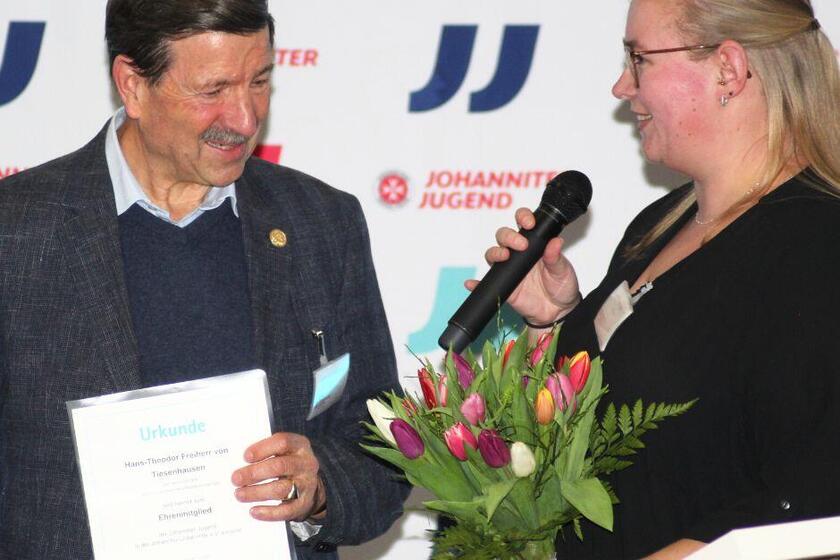 Hans-Theodor von Tiesenhausen hält eine Urkunde in der Hand, rechts neben ihm steht eine Jugendgruppenleiterin mit Mirkofon in der Hand.