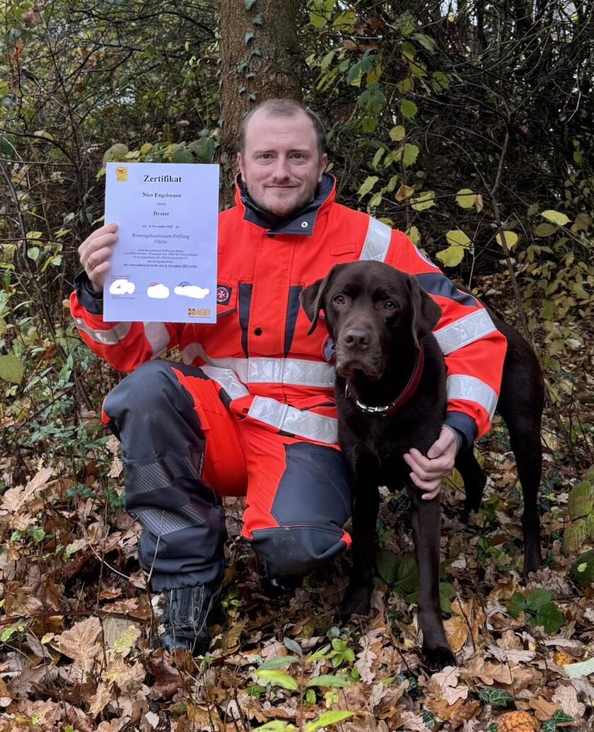 Ein Mitglied der Rettungshundestaffel in Einsatzkleidung kniet neben seinem Labrador Dexter und hält eine Urkunde in die Kamera.