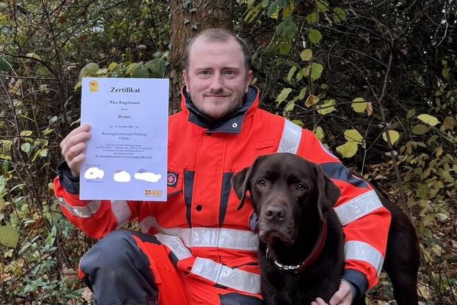 Ein Mitglied der Rettungshundestaffel in Einsatzkleidung kniet neben seinem Labrador Dexter und hält eine Urkunde in die Kamera.