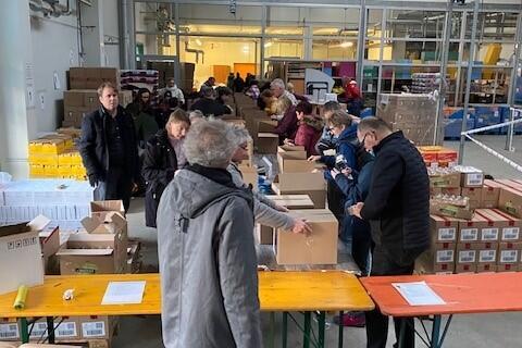 Zahlreiche Ehrenamtliche stehen an langen Tischen in einer großen Halle und packen Kartons mit Hilfsgütern für die Ukraine. Im Vordergrund packen Freiwillige die Pakete, rundherum stapeln sich Kisten mit Lebensmitteln und Hygieneartikeln. Das emsige Treiben zeigt das Engagement bei der gemeinsamen Packaktion von Johannitern, Rotary und Inner Wheel für die Ukraine-Hilfe.