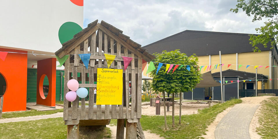 Eine Kinderholzhütte auf Stelzen mit einem gelben Pappschild auf dem "Sommerfest" steht. Dahinter einer Kita.