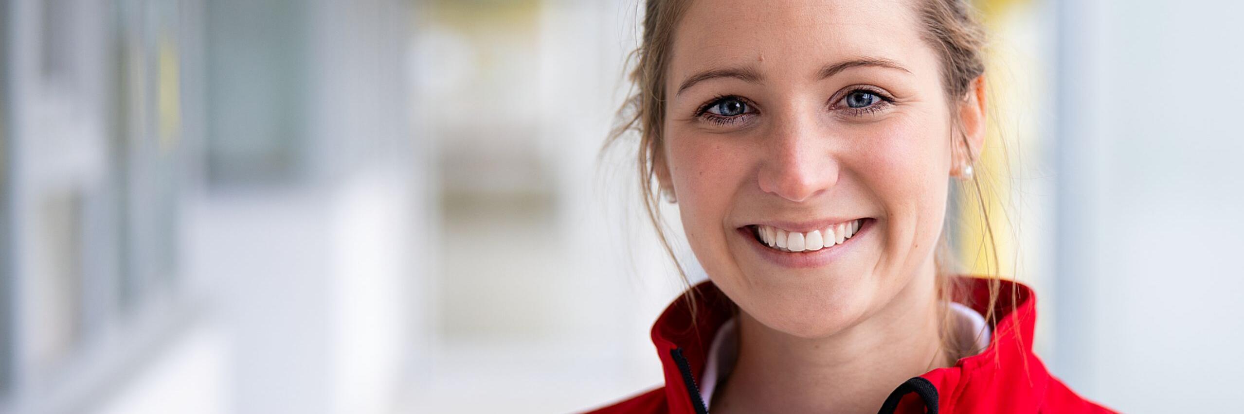 Person in roter Einsatzjacke lächelt in die Kamera, Innenbereich, unscharfer Hintergrund.