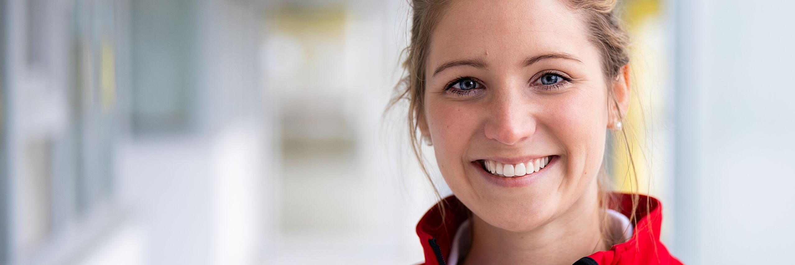 Person in roter Einsatzjacke lächelt in die Kamera, Innenbereich, unscharfer Hintergrund.
