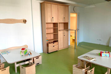 Grüner Fußboden, kleine Tische mit kleinen Stühlen inkl. Armlehnen. Auch ein Regal zum Aufbewahren von Spielsachen ist zu sehen.