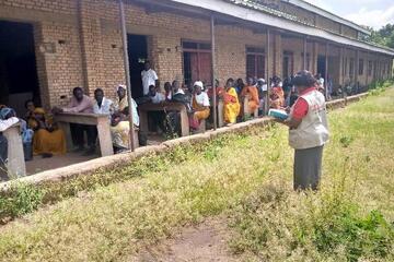 Eine Frau steht im Vordergrund und hält ein Buch in der Hand. Im Hintergrund sitzen Frauen und Männer an Tischen und schauen die Rednerin aufmerksam an