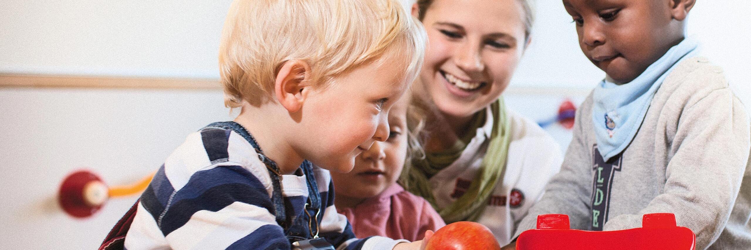 Eine Betreuungskraft der Johanniter sitzt mit drei Kindern am Tisch und spielt gemeinsam mit ihnen.