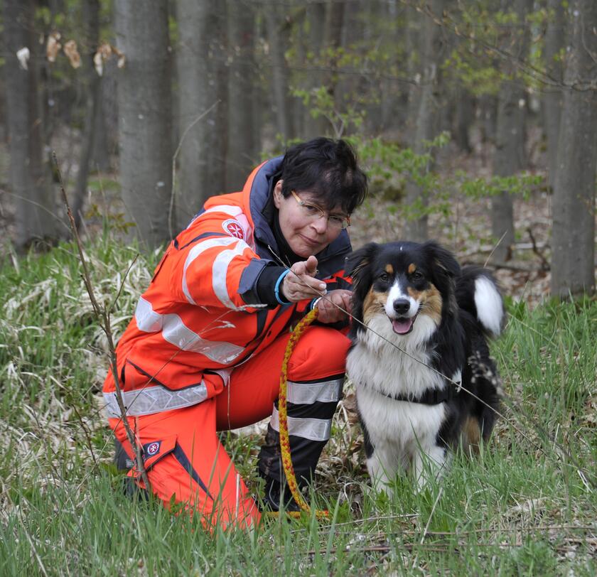 Eine Johanniter-Rettungshundeführerin hockt in leuchtender Einsatzkleidung im Wald neben ihrem weiß-braun-schwarzen Rettungshund. Beide lächeln in die Kamera und sind bereit für den nächsten Einsatz.