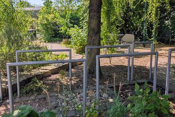 Blick in einen Garten, im Vordergrund Haltestangen links und rechts von einem Weg, der aus unterschiedlichem Bodenmaterial besteht 