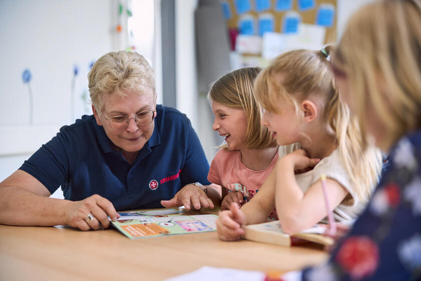 Johanniter-Mitarbeiterin liest mit Kindern ein Erste-Hilfe-Heft in fröhlicher Runde