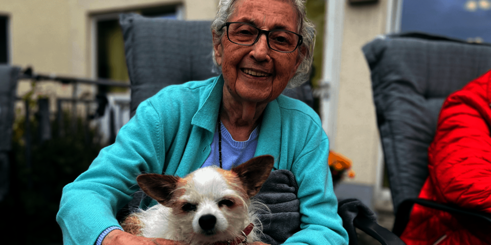 weißhaarige Frau in türkiser Jacke hält einen Hund auf dem Schoß