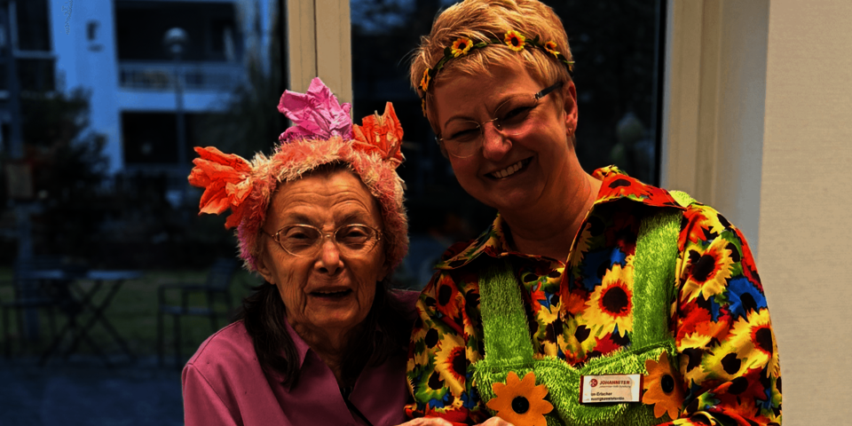 Zwei Frauen in bunten Kostümen lächeln in die Kamera, eine trägt Blumenkranz im Haar.