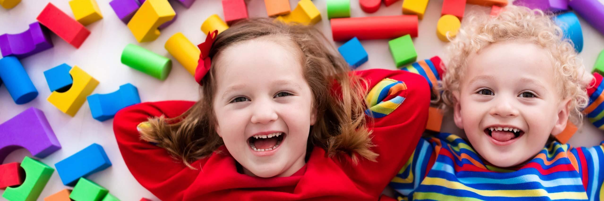 Zwei lachende Kinder liegen auf dem Boden zwischen bunten Spielbausteinen.