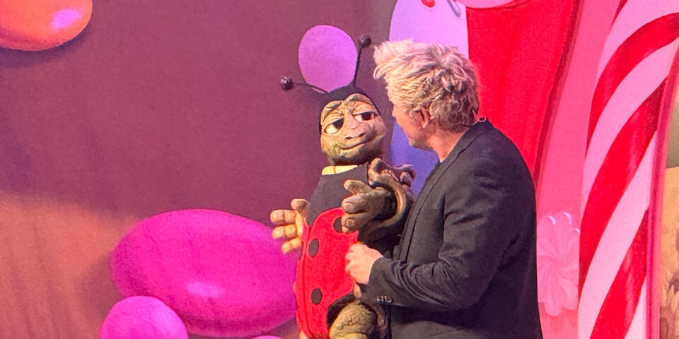 Ein blonder Mann mit einer Marienkäfer-Handpuppe auf einer bunten hell beleuchteten Bühne