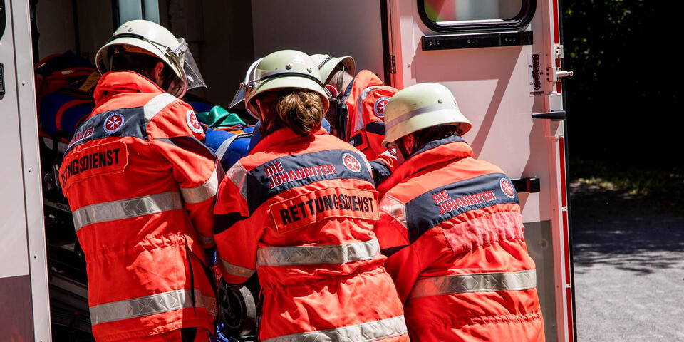 Rettungshelfer*innen heben gemeinsam eine verletzte Person auf einer Trage in den Rettungswagen.