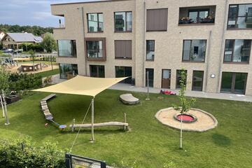 Die Außenansicht der Kita Kunterbunt mit begrünten Flächen, einem Sonnensegel und dem Ausblick auf einen Spielplatz mit Sandkasten.