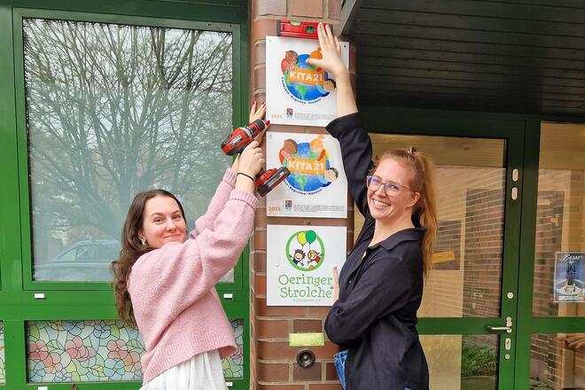 Stolz bringen Auszubildende Celina Schneider und Mitarbeiterin Jennifer Meyer das Auszeichnungsschild vor dem Eingang der Johanniter-Kita Oeringer Strolche an.
