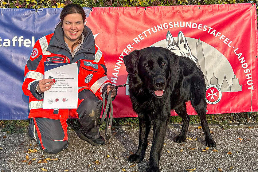 Person in Johanniter-Uniform kniet neben schwarzem Hund, hält Urkunde vor Rettungshundestaffel-Banner.
