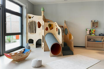 Ein Raum mit einer Holzwippe sowie einer kleinen Spielplattform mit Rutsche und Höhle.