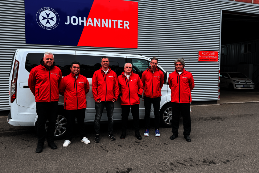 Sechs Männer in roten Jacken und dunklen Hosen stehen vor einem weißen Fahrzeug. Über einen an der Wand einer Fahrzeughalle ein Blau-rotes Schild mit dem Logo der Johanniter darauf.