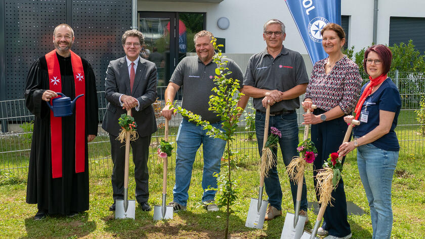 Ein Johanniter-Pfarrer und 5 weitere Johanniter stehen bei einer feierlichen Baumpflanzaktion vor einem Gebäude. Der Pfarrer hält eine Gießkanne in der Hand, die anderen halten jeweils einen geschmückten Spaten. Im Hintergrund ist eine Fahne, auf der "Johanniter" steht.