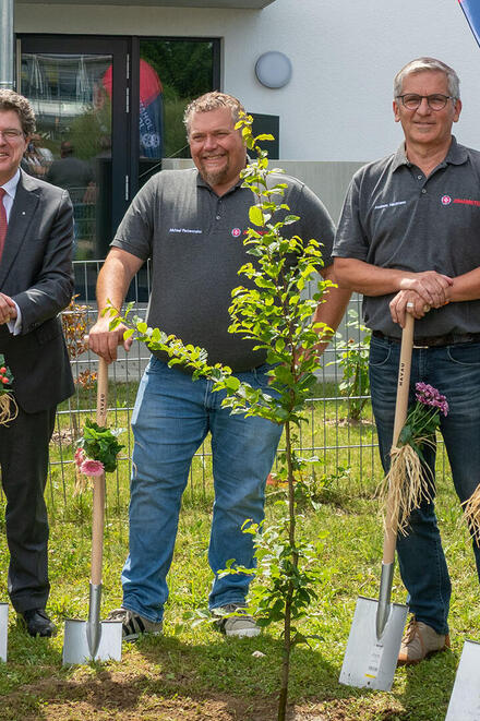 Ein Johanniter-Pfarrer und 5 weitere Johanniter stehen bei einer feierlichen Baumpflanzaktion vor einem Gebäude. Der Pfarrer hält eine Gießkanne in der Hand, die anderen halten jeweils einen geschmückten Spaten. Im Hintergrund ist eine Fahne, auf der "Johanniter" steht.
