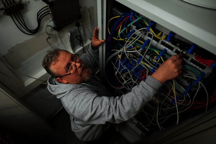 Ein Mann in grauer Arbeitskleidung arbeitet an einem Netzwerkschrank mit bunten Kabeln