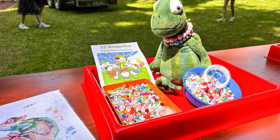 Ein Stoffchamäleon und kleine Kistchen mit bunten Perlen auf einem Tisch. Daneben ein Handzettel auf dem "32. Kinderfest" steht
