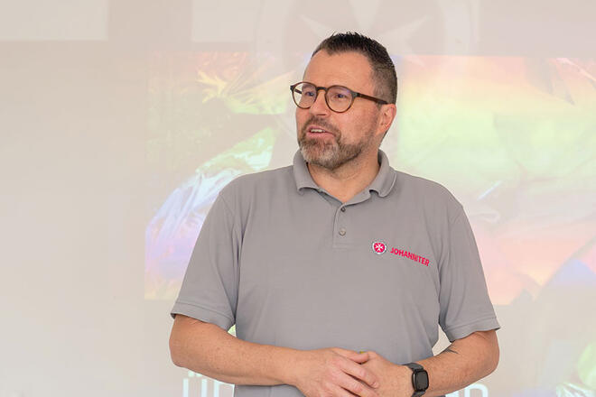 Ein Johanniter Mitarbeiter in Dienstkleidung steht vor einer weißen Wand.