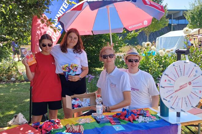 Ein bunter Stand der Johanniter-Jugend mit Drehrad, Flyern, Stiften, BonBons. Am Stand posieren vier Jugendliche in Johanniter-Jugend Klamotten. 