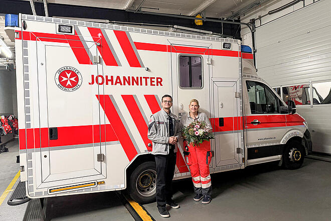 Ein Mann mit Johanniter-Jacke und eine Frau in Einsatzkleidung und mit einem Blumenstrauß in der Hand vor einem RTW
