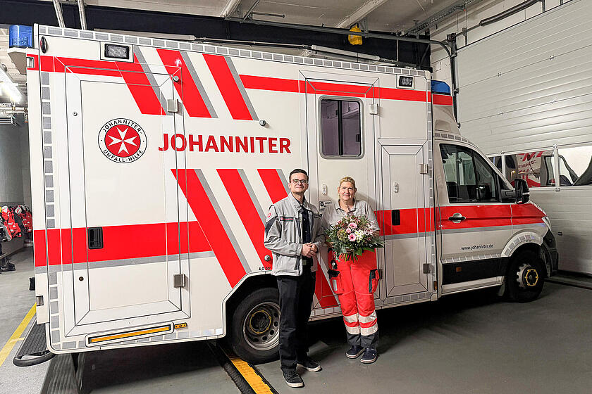 Ein Mann mit Johanniter-Jacke und eine Frau in Einsatzkleidung und mit einem Blumenstrauß in der Hand vor einem RTW