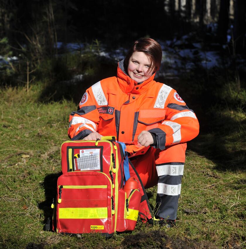 Eine Johanniter-Helferin sitzt in leuchtender Einsatzkleidung im Wald auf einer Wiese und lächelt in die Kamera. Vor ihr steht ein großer, auffälliger Notfallrucksack mit Reflexstreifen, der für den Rettungseinsatz bereit ist.
