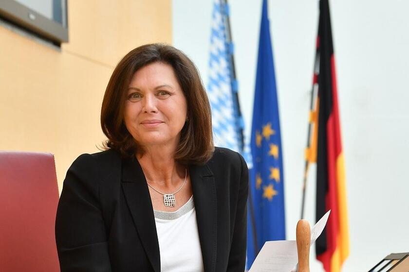 Frau sitzt mit Dokumenten am Rednerpult, hinter ihr Flaggen von Deutschland, Bayern und der EU.