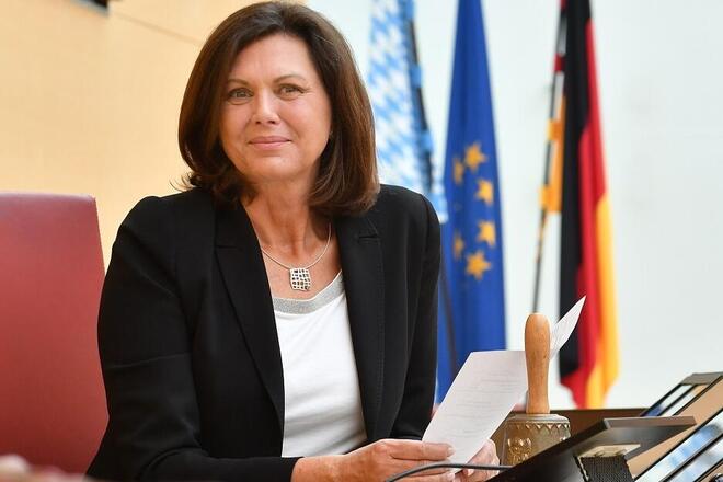 Frau sitzt mit Dokumenten am Rednerpult, hinter ihr Flaggen von Deutschland, Bayern und der EU.