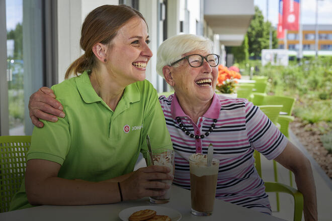 Eine Senioren legt den Arm um eine Pflegekraft der Johanniter, beide lächeln.