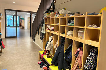 Eingangsbereich der Kita zeigt einen Flur mit Garderobe für Kinder, die als offenes Regal gebaut ist. Hierin befinden sich die Jacken, Schuhe und oberhalb Möglichkeiten für kleinere Dinge unterzubringen.