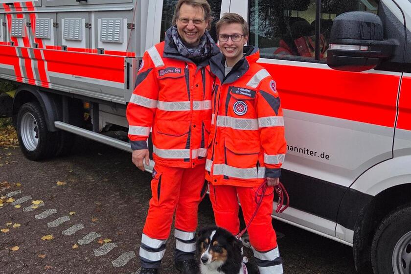 Ein Mann und eine Frau in Einsatzkleidung stehen zusammen mit einem Hund mit Kenndecke vor einem Einsatzfahrzeug.