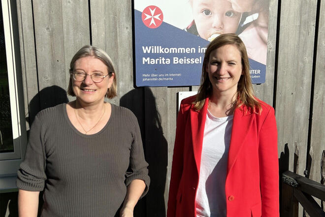 Zwei Frauen stehen vor einer Hauswand. Hinter ihnen hängt ein Schild auf dem steht „Willkommen im Marita Beissel Haus“.