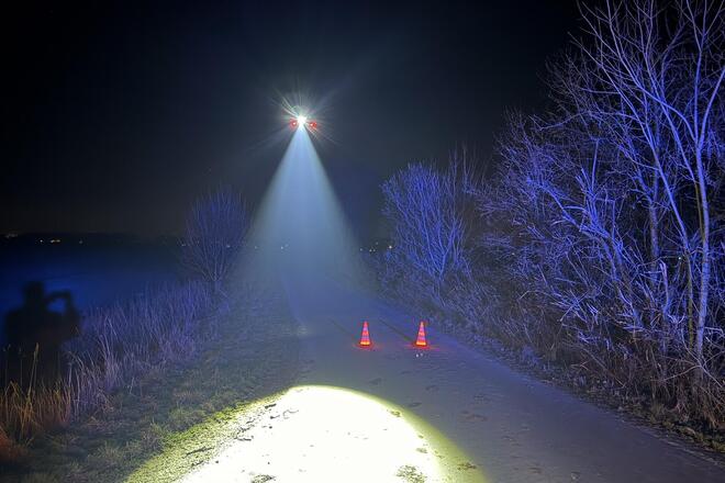Es ist tiefe Nacht. Eine fliegende Drohne beleuchtet einen Feldweg auf dem zwei Leitkegel stehen.
