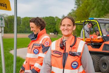 Zwei Johanniterinnen in orangefarbener Einsatzkleidung stehen im Freien, im Hintergrund Einsatzfahrzeug.