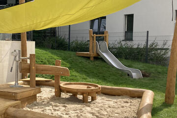 Wasserspielanlage, Sandkasten, Rutsche und gelbes Sonnensegel sind zu sehen. 