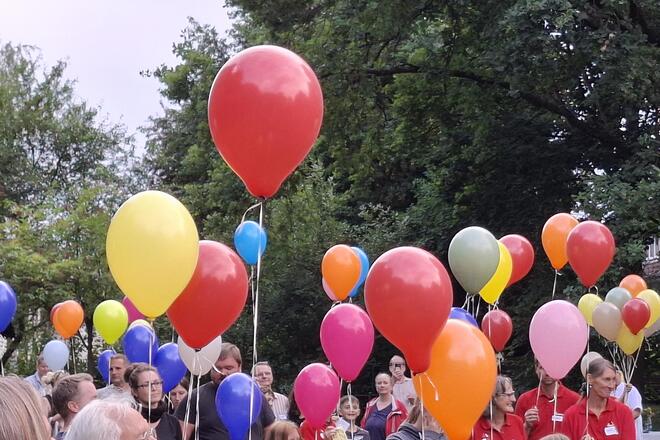 Viele Menschen im Freien halten bunte Luftballons in die Höhe, im Vordergrund sind Bäume zu sehen.