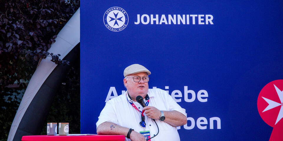 Mann mit Mikrofon und Namensschild spricht vor einem Johanniter‑Messestand zu den Anwesenden und verkündet die Gewinner.