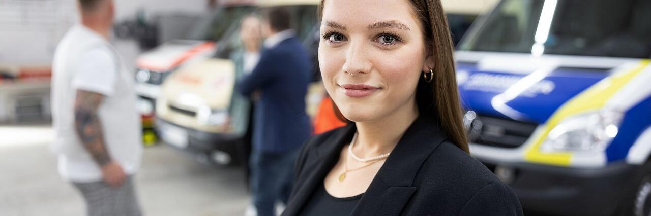 Junge Frau im schwarzen Blazer steht vor Einsatzfahrzeugen.