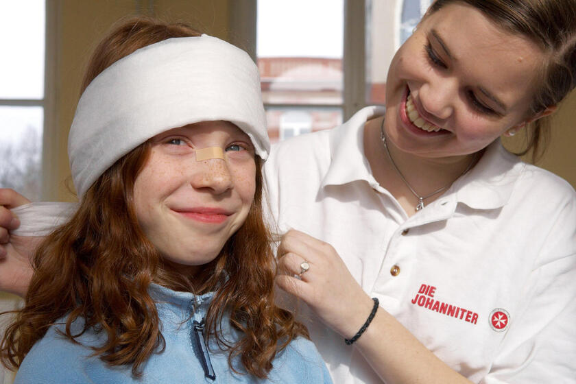 Eine Sanitäterin untersucht ein lächelndes Mädchen mit Kopfverband in einem Behandlungsraum.