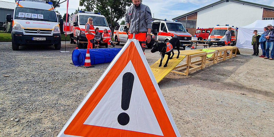 Ein Faltsignal mit der Aufschrift "Rettungshunde" steht auf einem Platz. Dahinter geht ein Hund, angeleitet von einem Mann über eine Holzbrücke. Im Hintergrund stehen Einsatzfahrzeuge