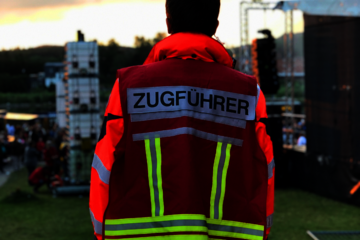 Ein ehrenamtlicher Helfer in Rückenansicht blickt auf ein Festivalgeschehen vor ihm an einem See. Er trägt eine Weste, die ihn als Zugführer ausweist.