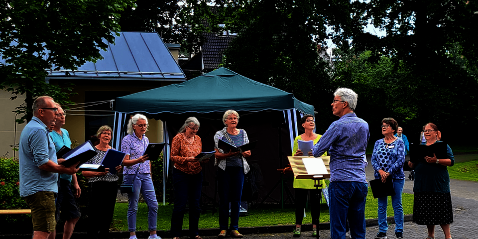 Eine Gruppe steht singend im Freien. Eine Person dirigiert.